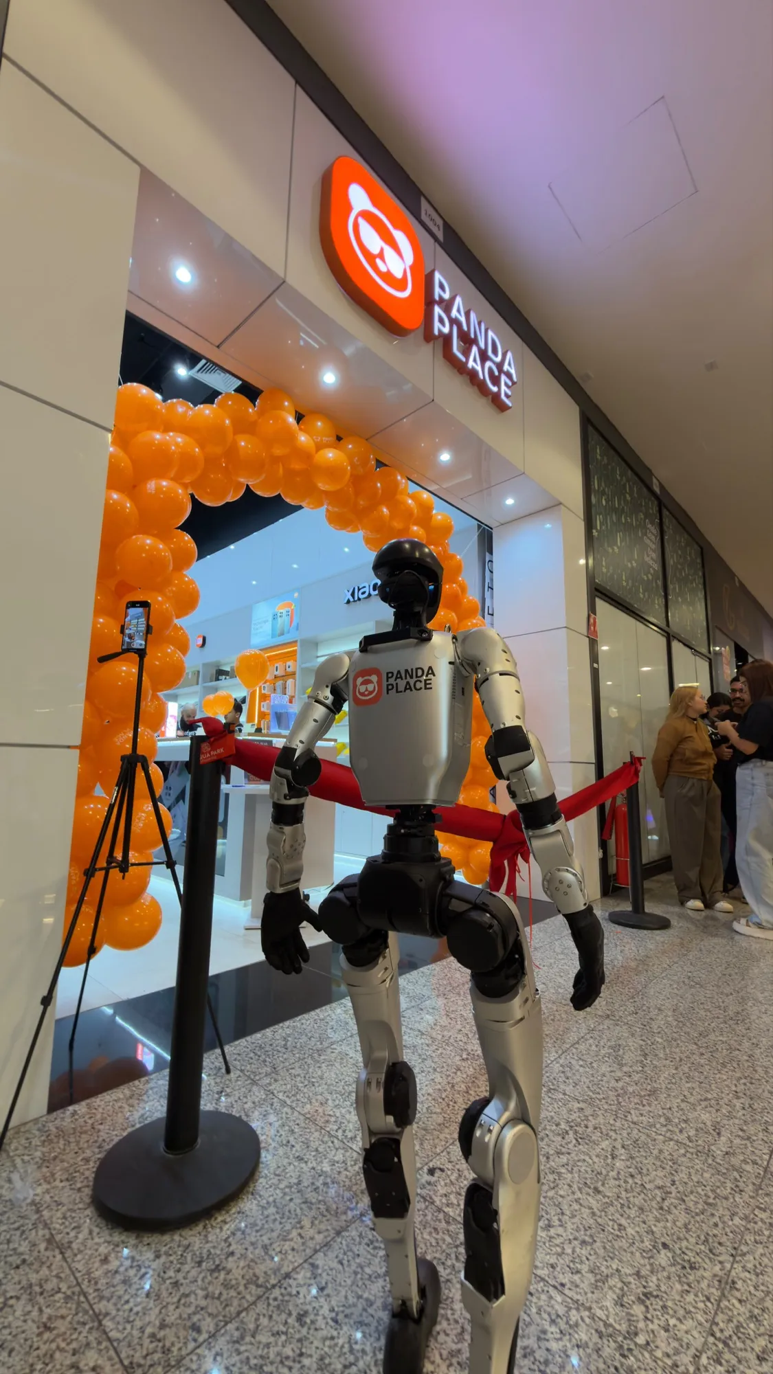 Inauguração de uma loja Panda Place, com arco de balões laranja e um robô humanoide na entrada